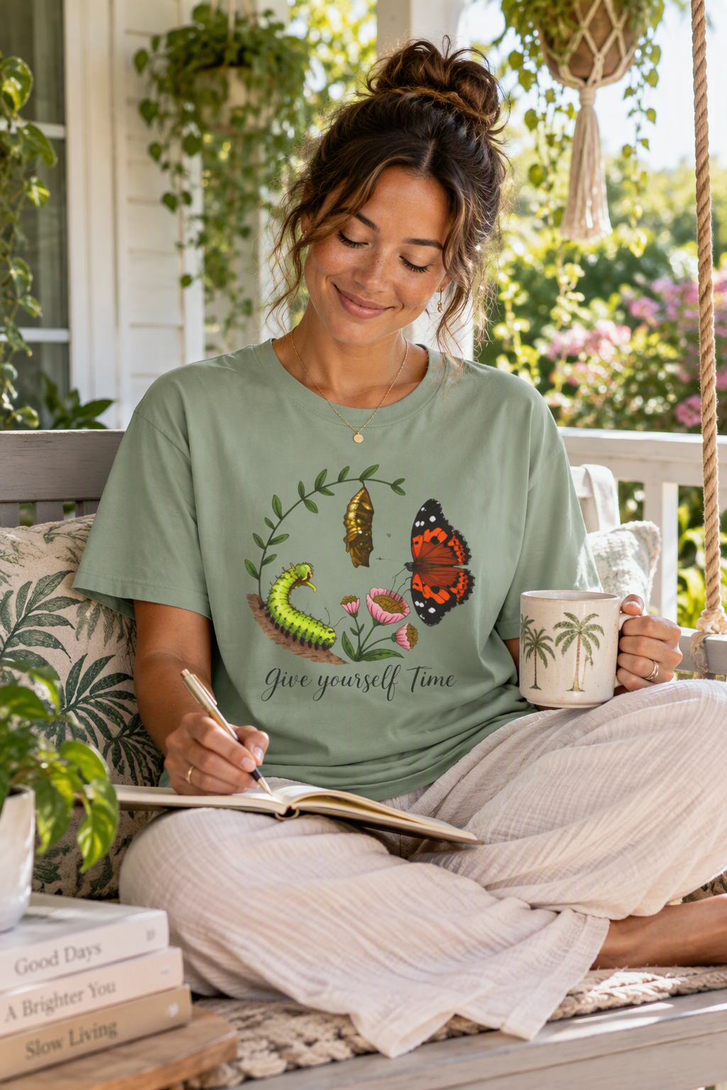 Woman journaling on a porch wearing the sage Give Yourself Time butterfly transformation tee